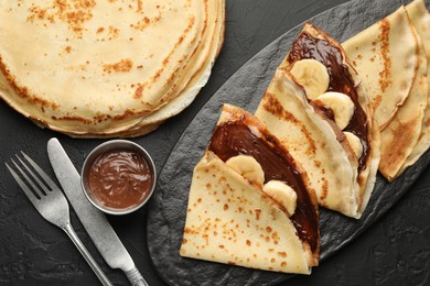 Photo of Tasty crepes with chocolate butter, banana and cutlery on black table, flat lay