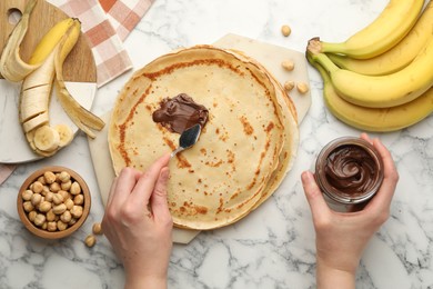 Photo of Woman spreading tasty chocolate butter onto crepe at white marble table, top view