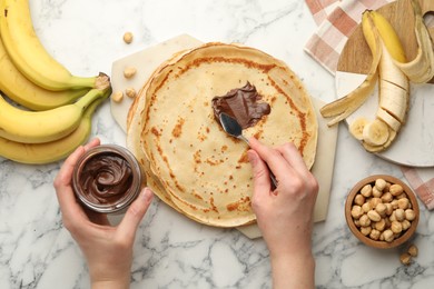 Photo of Woman spreading tasty chocolate butter onto crepe at white marble table, top view