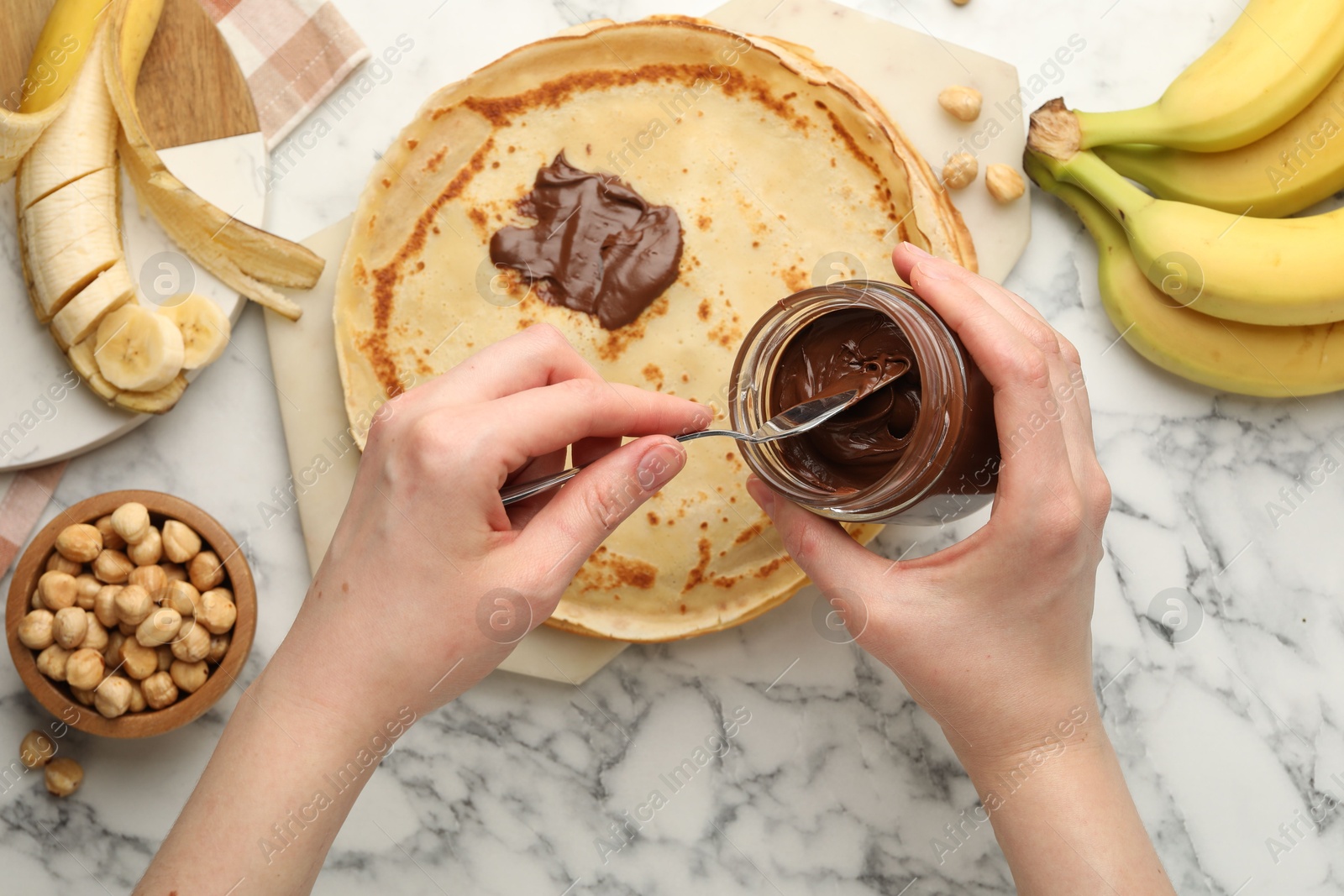 Photo of Woman spreading tasty chocolate butter onto crepe at white marble table, top view