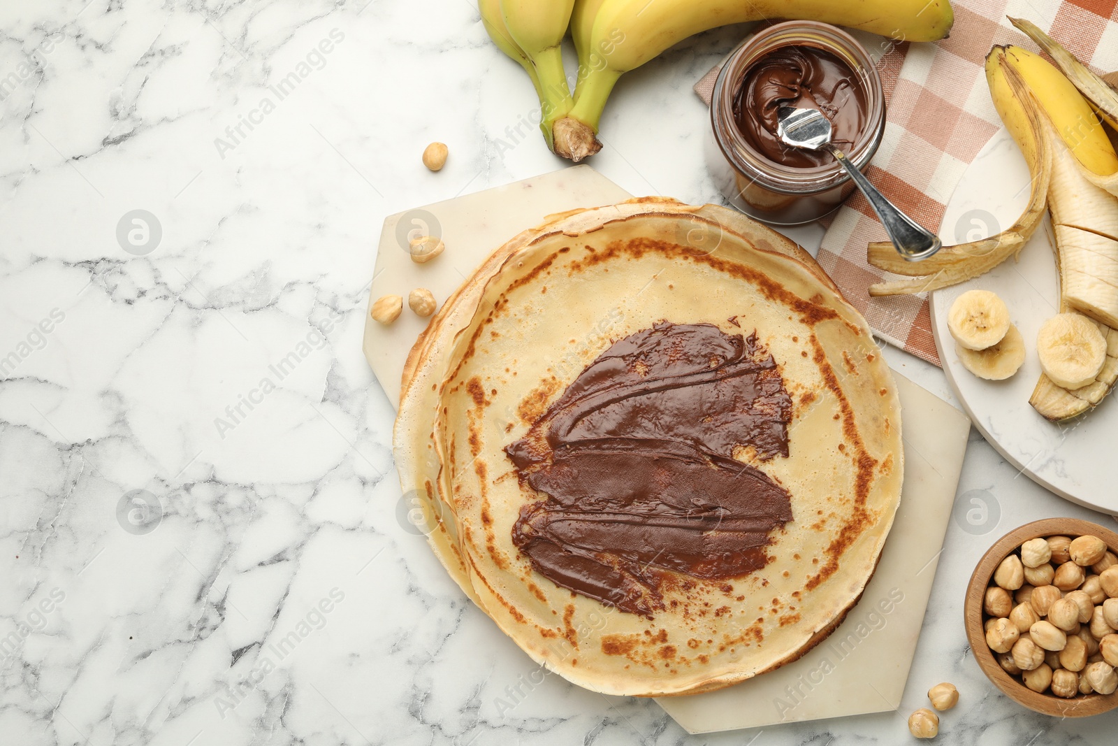 Photo of Tasty crepes with chocolate butter, bananas and hazelnuts on white marble table, flat lay. Space for text