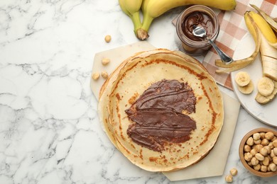 Tasty crepes with chocolate butter, bananas and hazelnuts on white marble table, flat lay. Space for text