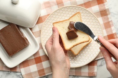 Photo of Woman spreading tasty chocolate butter onto bread at white marble table, top view