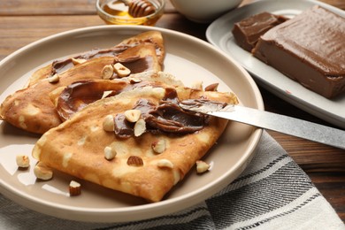 Photo of Tasty crepes with chocolate butter, nuts, honey and knife on wooden table, closeup