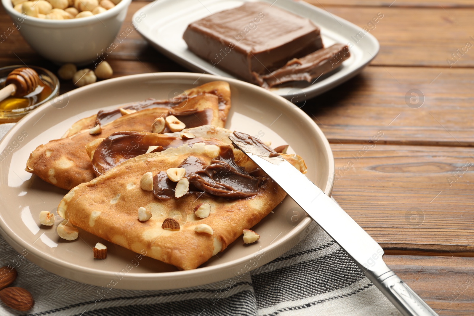 Photo of Tasty crepes with chocolate butter, nuts, honey and knife on wooden table, closeup. Space for text