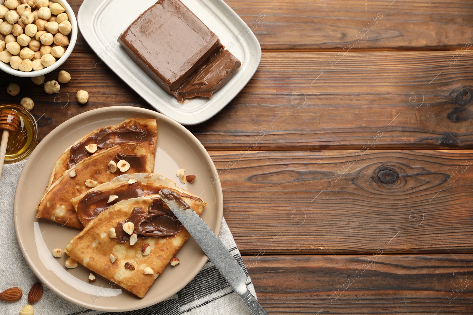 Photo of Tasty crepes with chocolate butter, nuts, honey and knife on wooden table, flat lay. Space for text