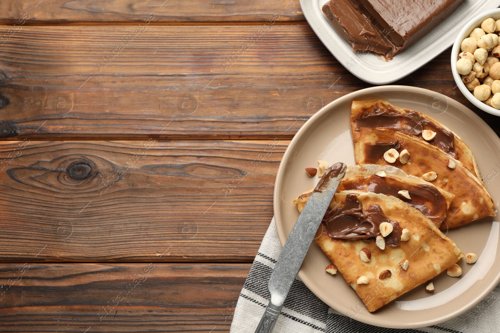 Photo of Tasty crepes with chocolate butter, nuts and knife on wooden table, flat lay. Space for text