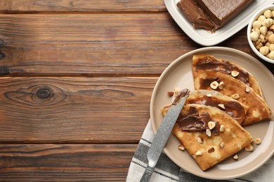 Photo of Tasty crepes with chocolate butter, nuts and knife on wooden table, flat lay. Space for text