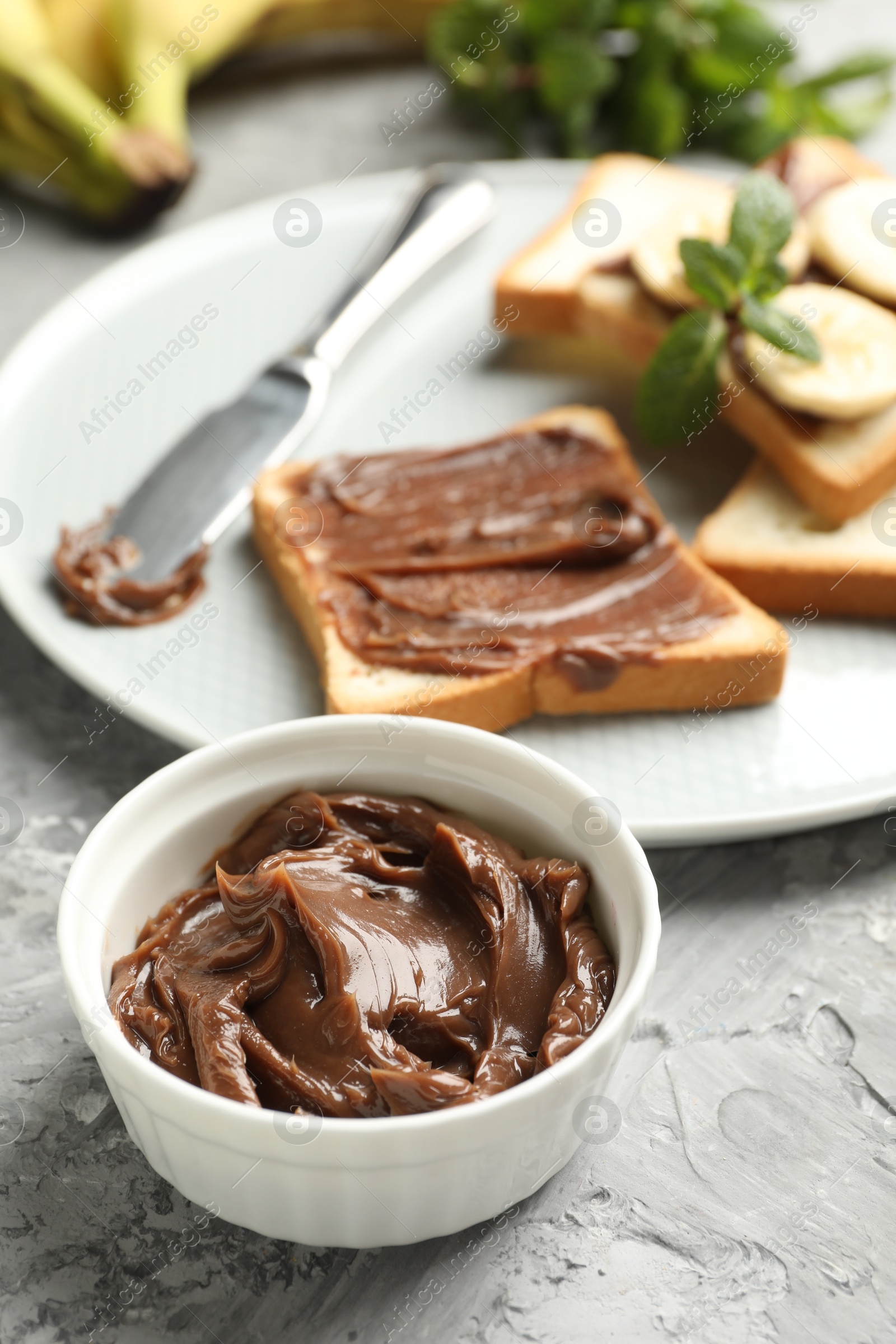 Photo of Tasty chocolate butter in bowl, sandwiches, bananas, mint and knife on grey table, closeup