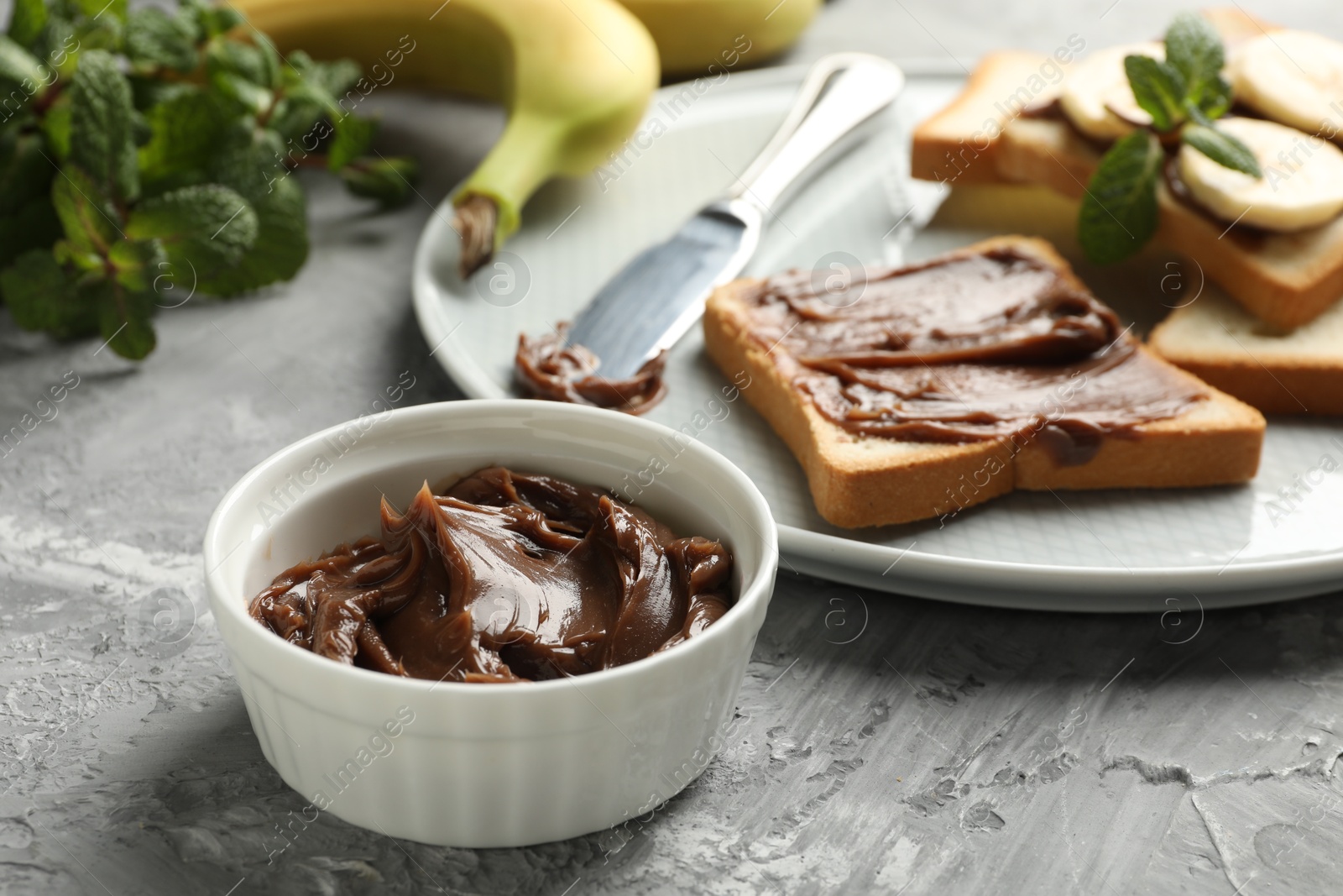 Photo of Tasty chocolate butter in bowl, sandwiches, bananas, mint and knife on grey table, closeup