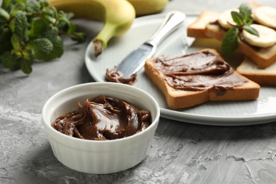 Tasty chocolate butter in bowl, sandwiches, bananas, mint and knife on grey table, closeup