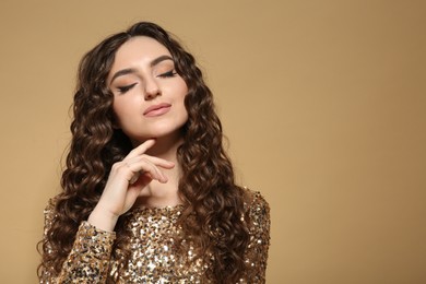 Photo of Beautiful young woman with long curly brown hair in golden sequin dress on beige background, space for text