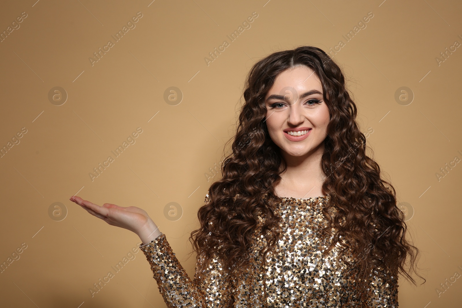 Photo of Beautiful young woman with long curly brown hair in golden sequin dress on beige background, space for text