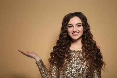 Photo of Beautiful young woman with long curly brown hair in golden sequin dress on beige background, space for text