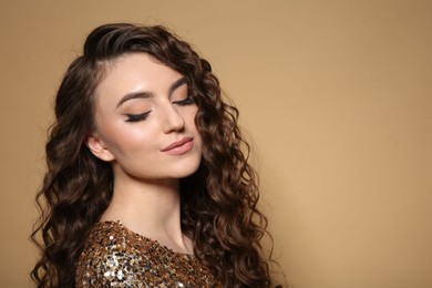 Photo of Beautiful young woman with long curly brown hair in golden sequin dress on beige background, space for text