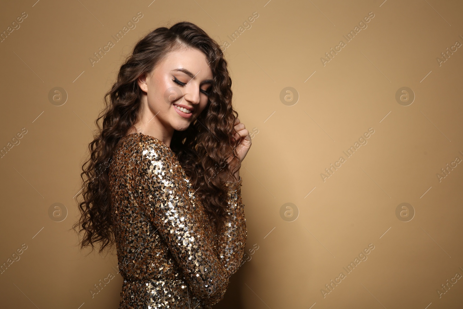 Photo of Beautiful young woman with long curly brown hair in golden sequin dress on beige background, space for text