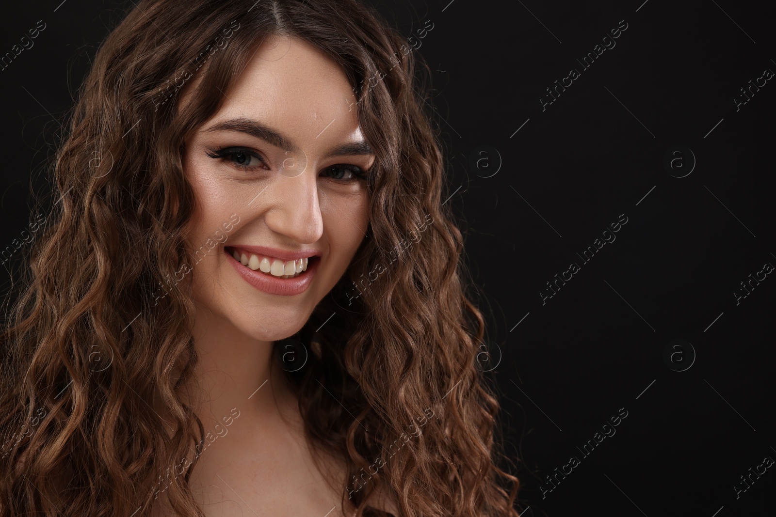 Photo of Beautiful young woman with long curly brown hair on black background, closeup. Space for text