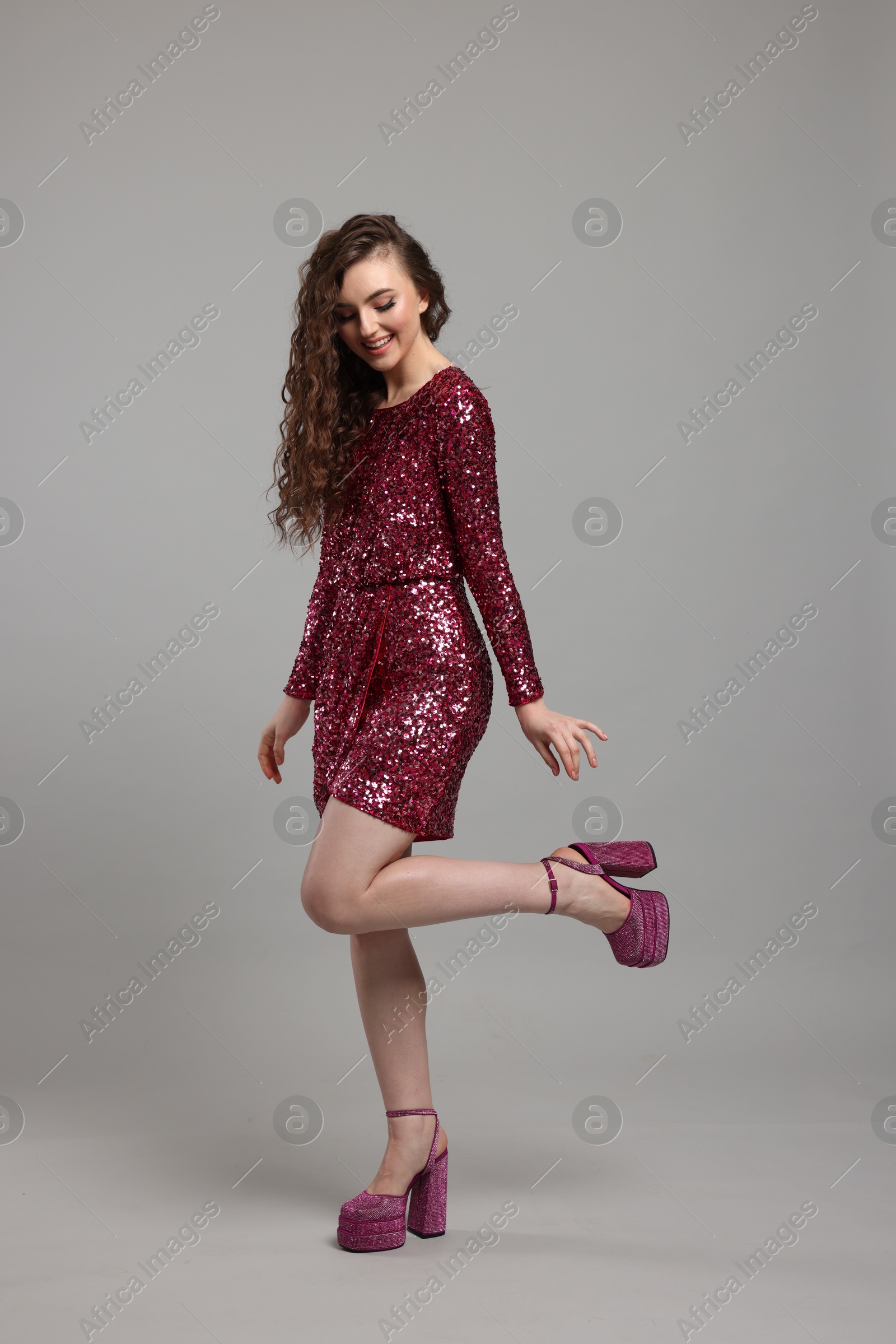 Photo of Beautiful young woman with long curly brown hair in pink sequin dress on grey background