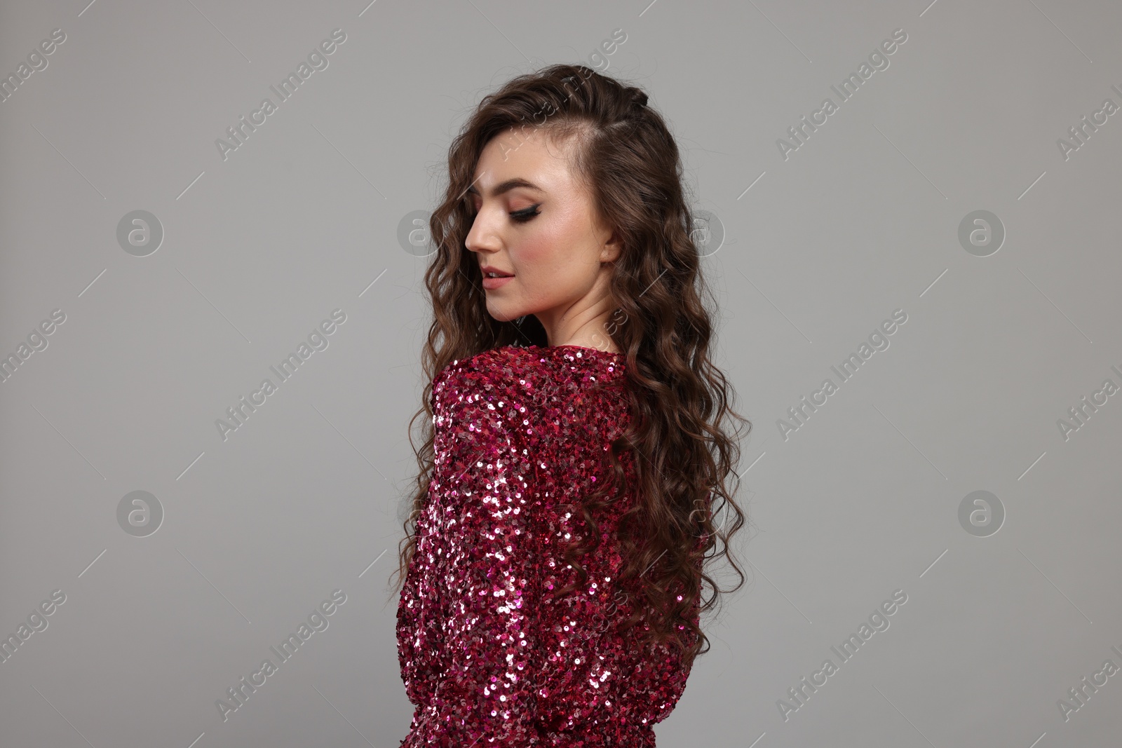 Photo of Beautiful young woman with long curly brown hair in pink sequin dress on grey background