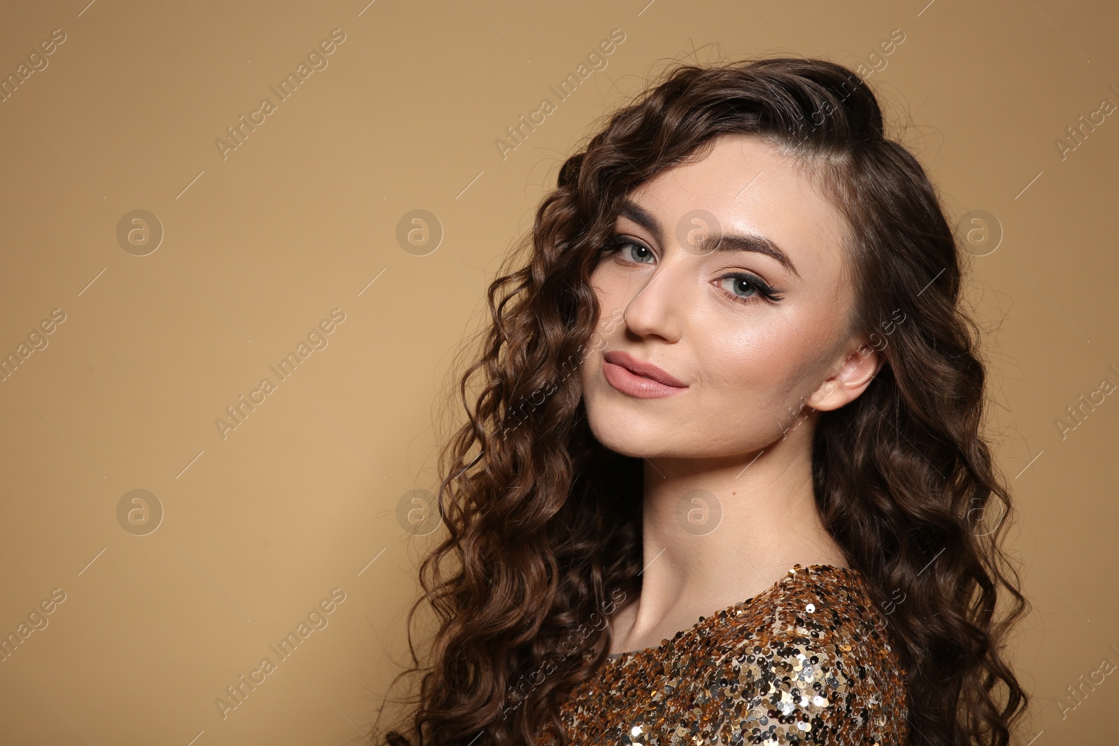 Photo of Beautiful young woman with long curly brown hair in golden sequin dress on beige background. Space for text