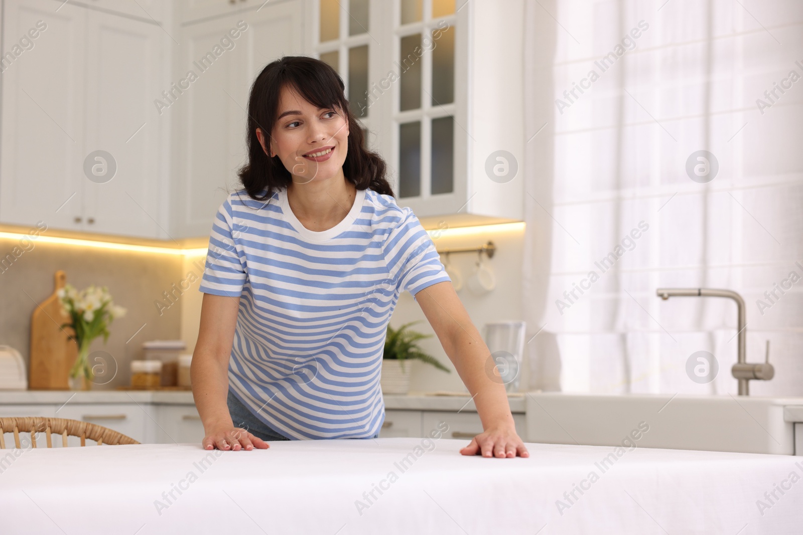 Photo of Young woman putting white tablecloth on table in kitchen