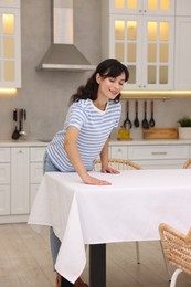 Photo of Young woman putting white tablecloth on table in kitchen