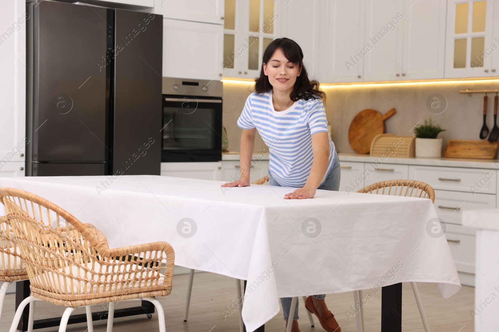 Photo of Young woman putting white tablecloth on table in kitchen