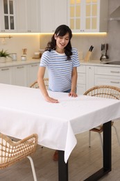 Photo of Young woman putting white tablecloth on table in kitchen