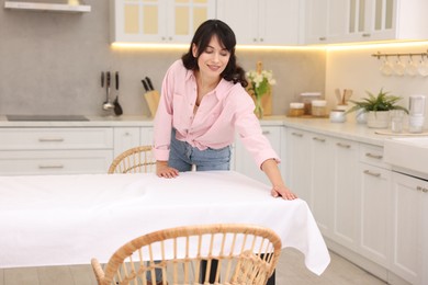 Photo of Young woman putting white tablecloth on table in kitchen