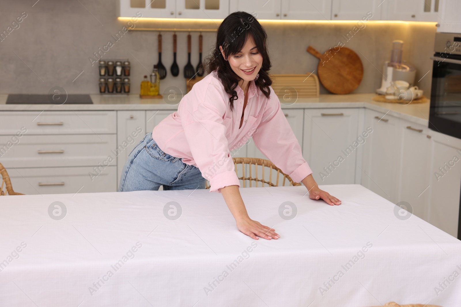 Photo of Young woman putting white tablecloth on table in kitchen