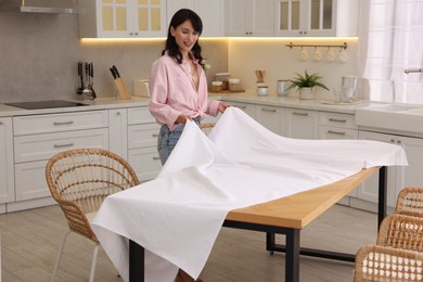 Photo of Young woman putting white tablecloth on table in kitchen