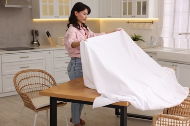 Photo of Young woman putting white tablecloth on table in kitchen