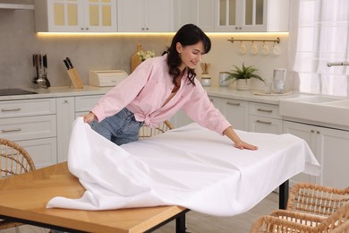 Photo of Young woman putting white tablecloth on table in kitchen