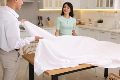 Photo of Couple putting white tablecloth on table in kitchen, selective focus