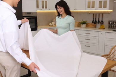 Photo of Couple putting white tablecloth on table in kitchen