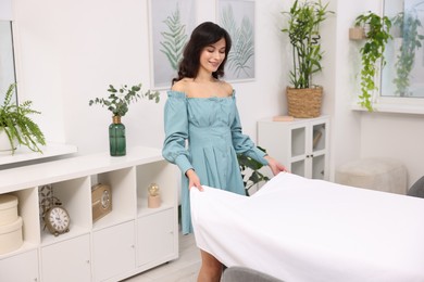 Photo of Young woman putting white tablecloth on table at home