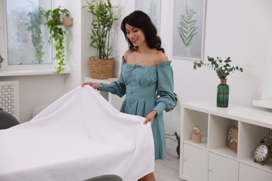 Photo of Young woman putting white tablecloth on table at home