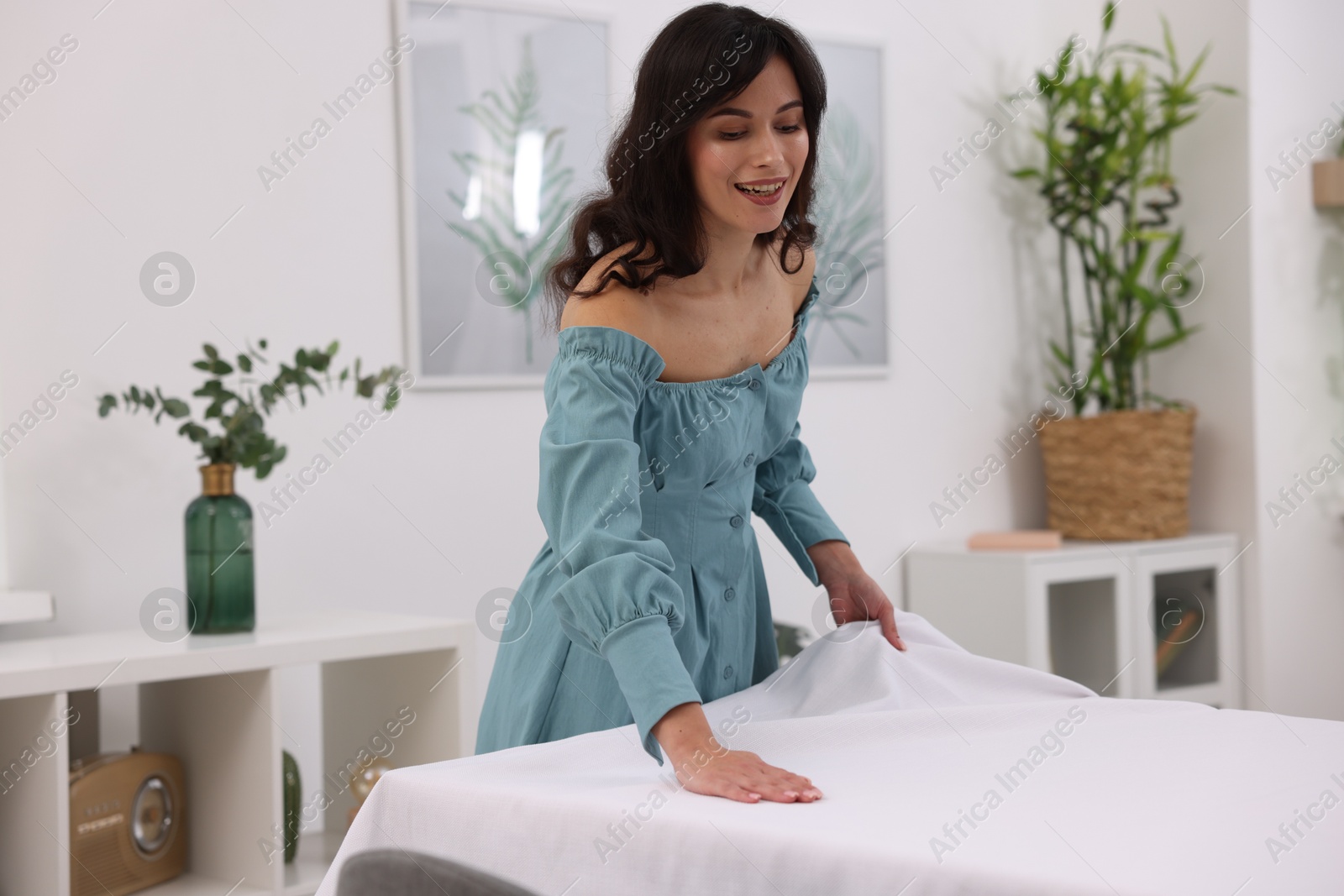 Photo of Young woman putting white tablecloth on table at home