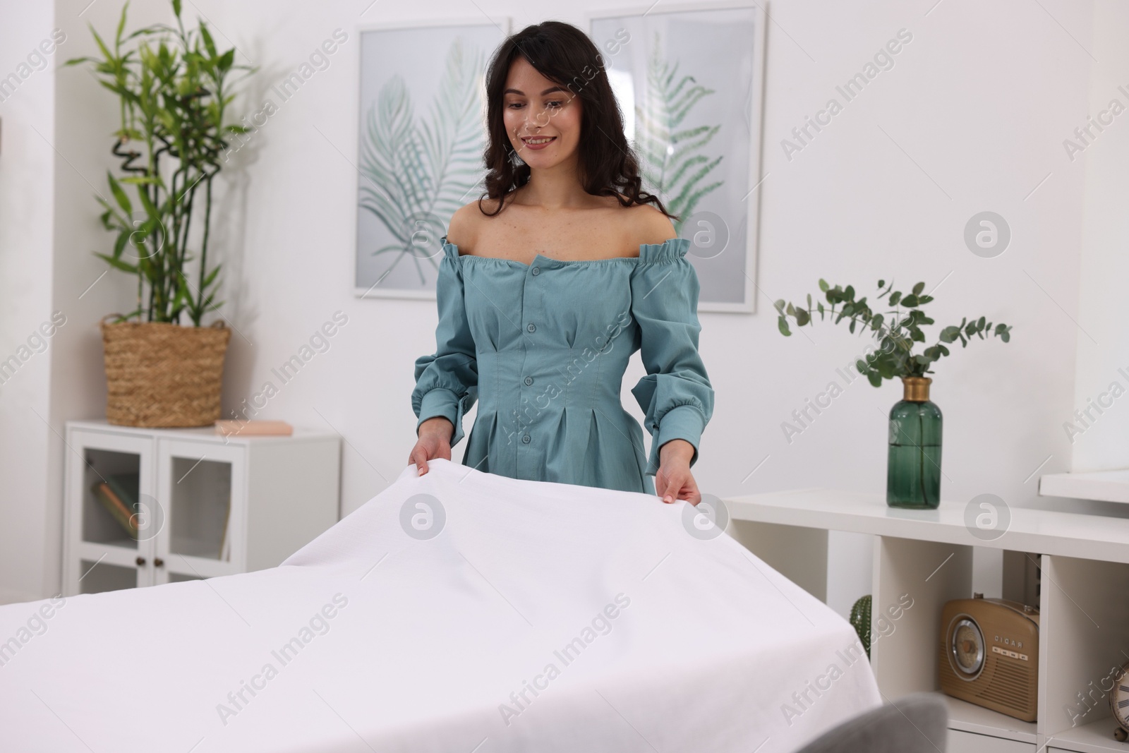 Photo of Young woman putting white tablecloth on table at home