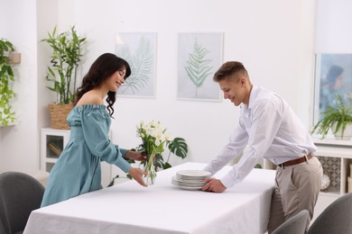 Photo of Couple putting plates and vase with flowers on table with tablecloth at home