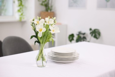 Photo of Plates and flowers in vase on table with white tablecloth indoors