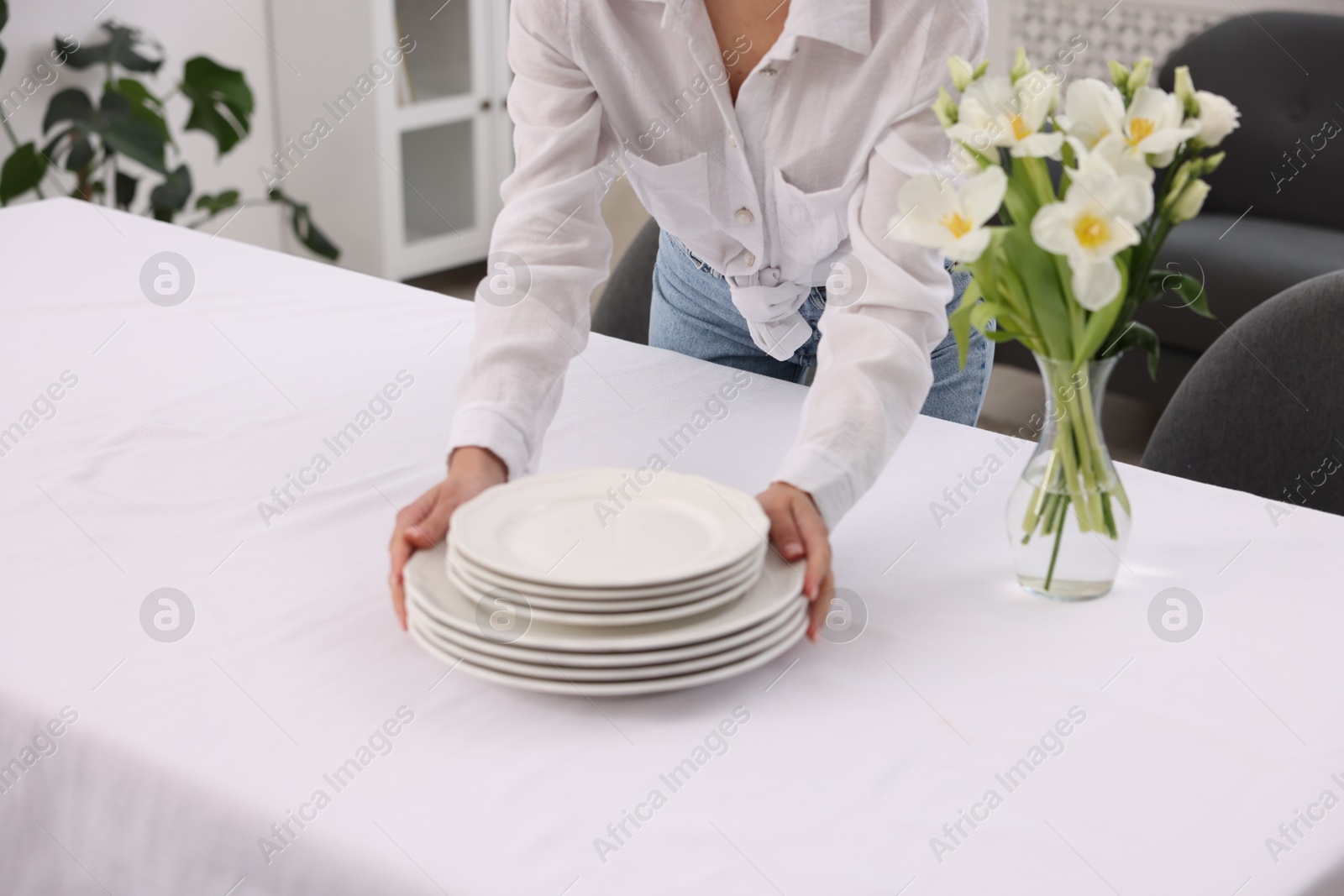 Photo of Young woman putting plates on table with white tablecloth at home, closeup