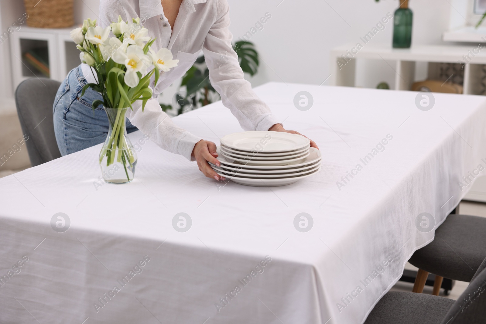 Photo of Young woman putting plates on table with white tablecloth at home, closeup