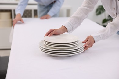 Photo of Couple putting plates on table with white tablecloth at home, closeup