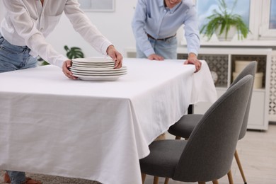 Photo of Couple putting plates on table with white tablecloth at home, closeup