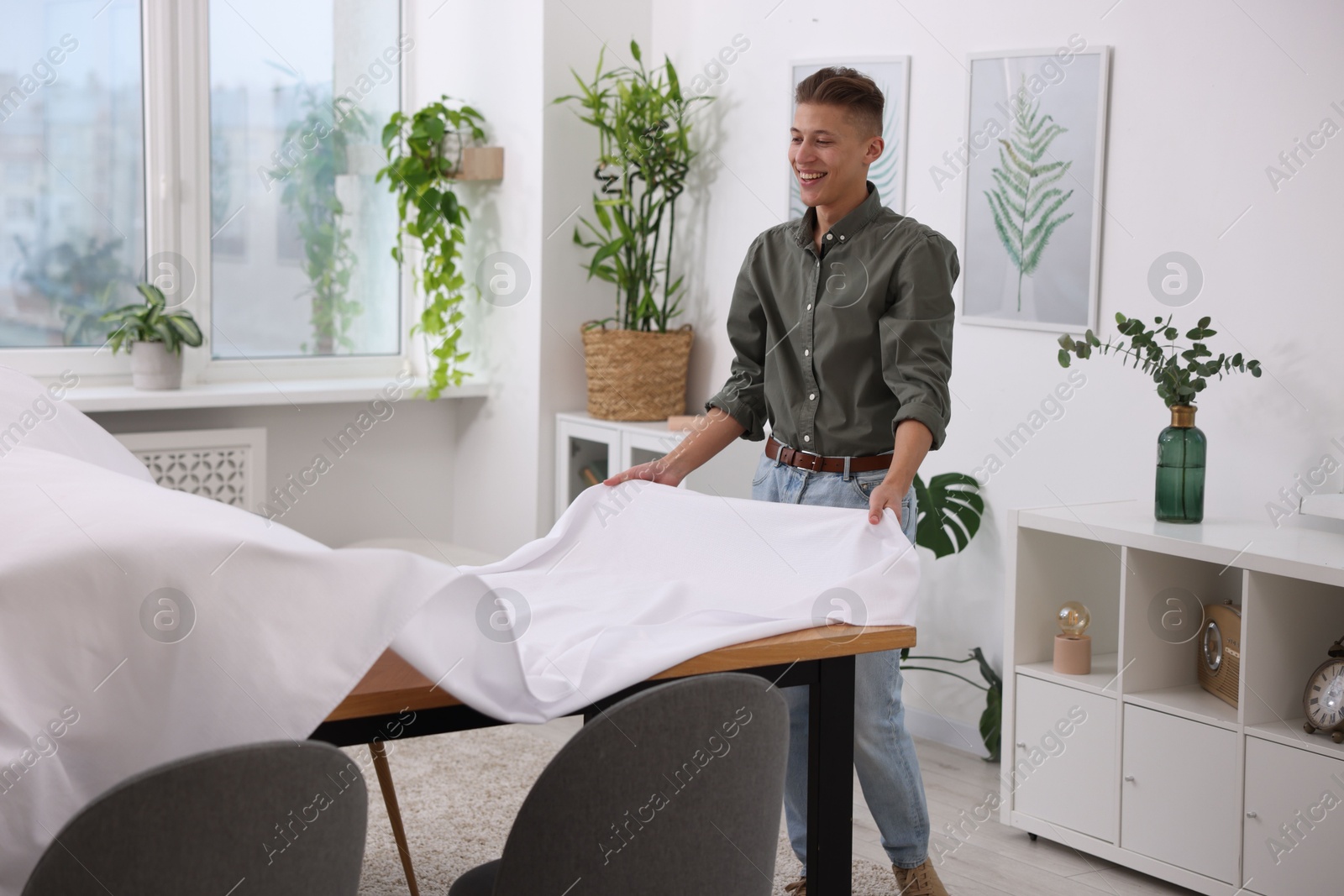 Photo of Young man putting white tablecloth on table at home