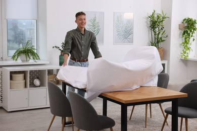 Photo of Young man putting white tablecloth on table at home