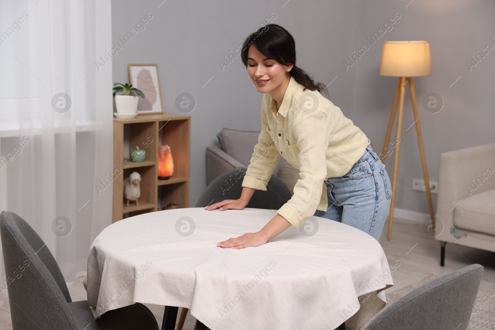 Photo of Young woman putting white tablecloth on table at home