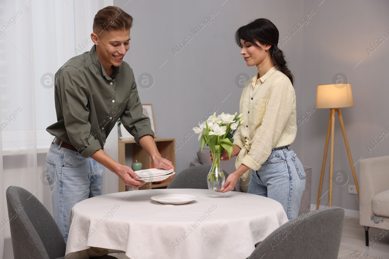 Photo of Couple putting plates and vase with flowers on table with white tablecloth at home