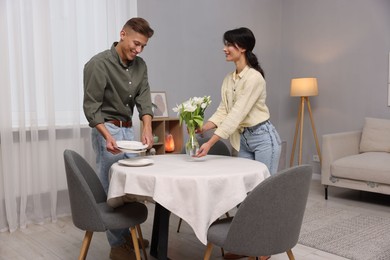 Photo of Couple putting plates and vase with flowers on table with white tablecloth at home